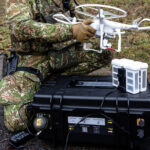 Energy Case noir fermé de B&W International au premier plan - en arrière-plan, un soldat en camouflage avec un drone blanc en mission militaire extérieure.