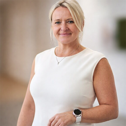 Eine Frau mit zurückgebundenen blonden Haaren, die ein ärmelloses weißes Oberteil und eine Smartwatch trägt, steht in einem Haus und lächelt in die Kamera, die neben ihrem B&W International-Koffer steht.